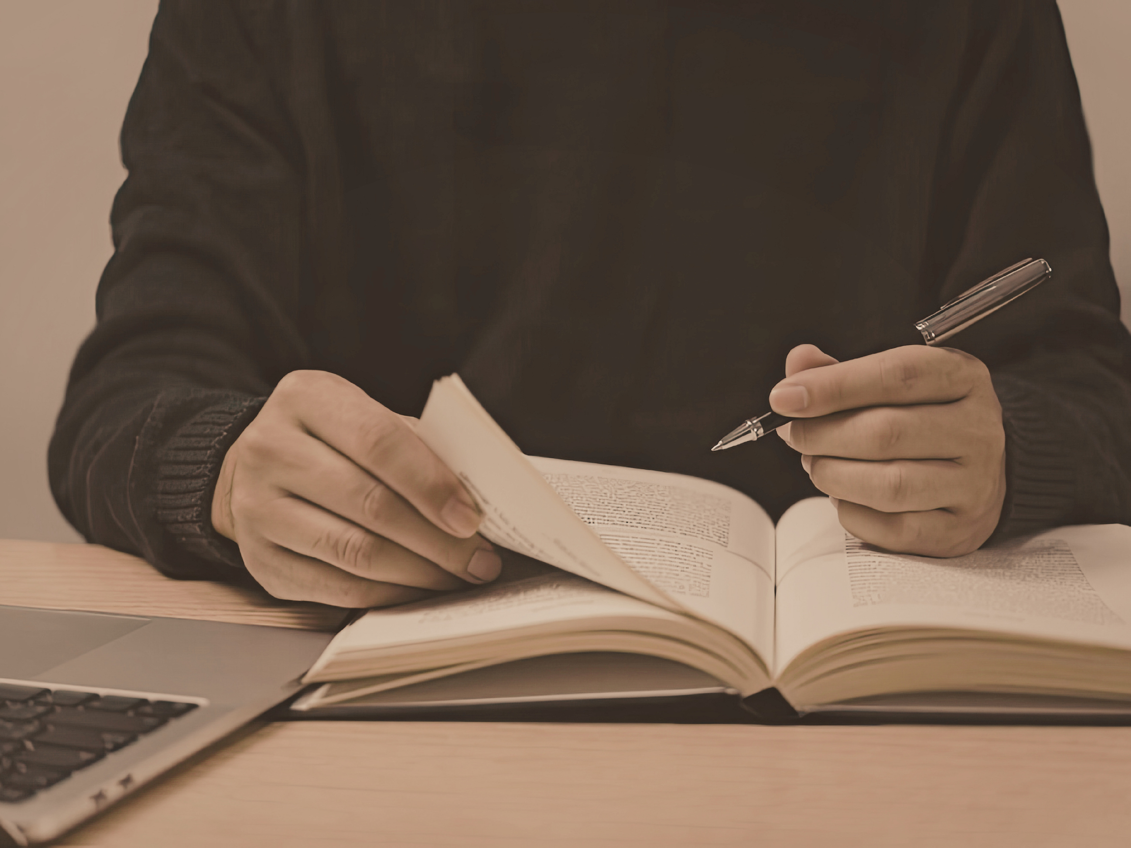 Une personne lisant un livre et écrivant, un ordinateur portable à côté